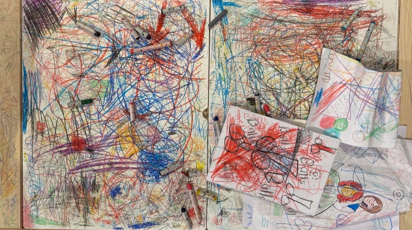 Chaotische bunte Wachsmalstift-Kritzeleien eines Kindes auf Tafeln und losem Papier, mit verstreuten Stiften auf einem Holztisch